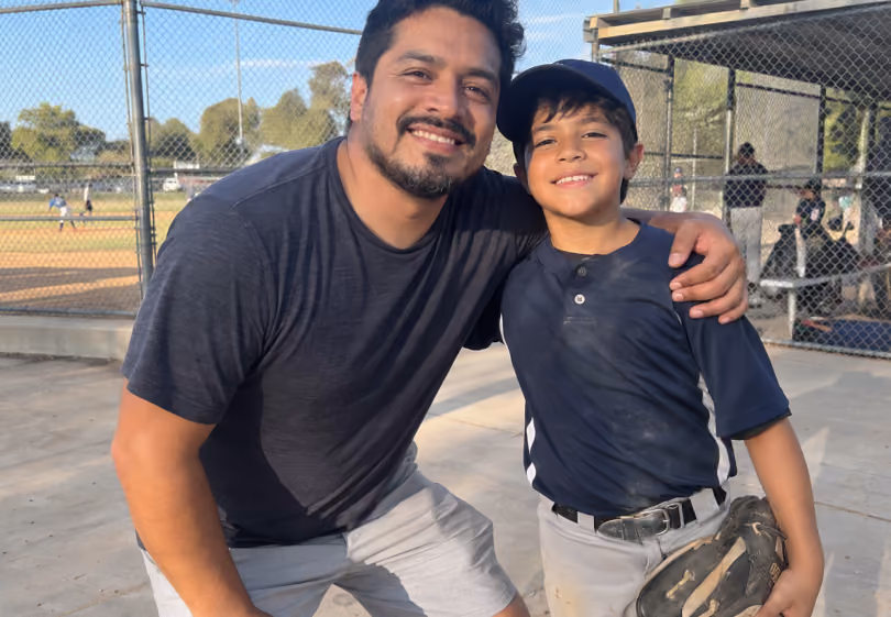 Dad and son in a baseball uniform smiling together at the ballfield