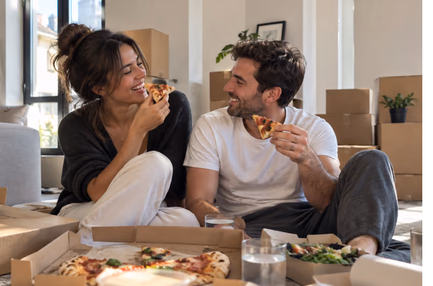 Couple eating pizza on the floor surrounded by moving boxes in their new home