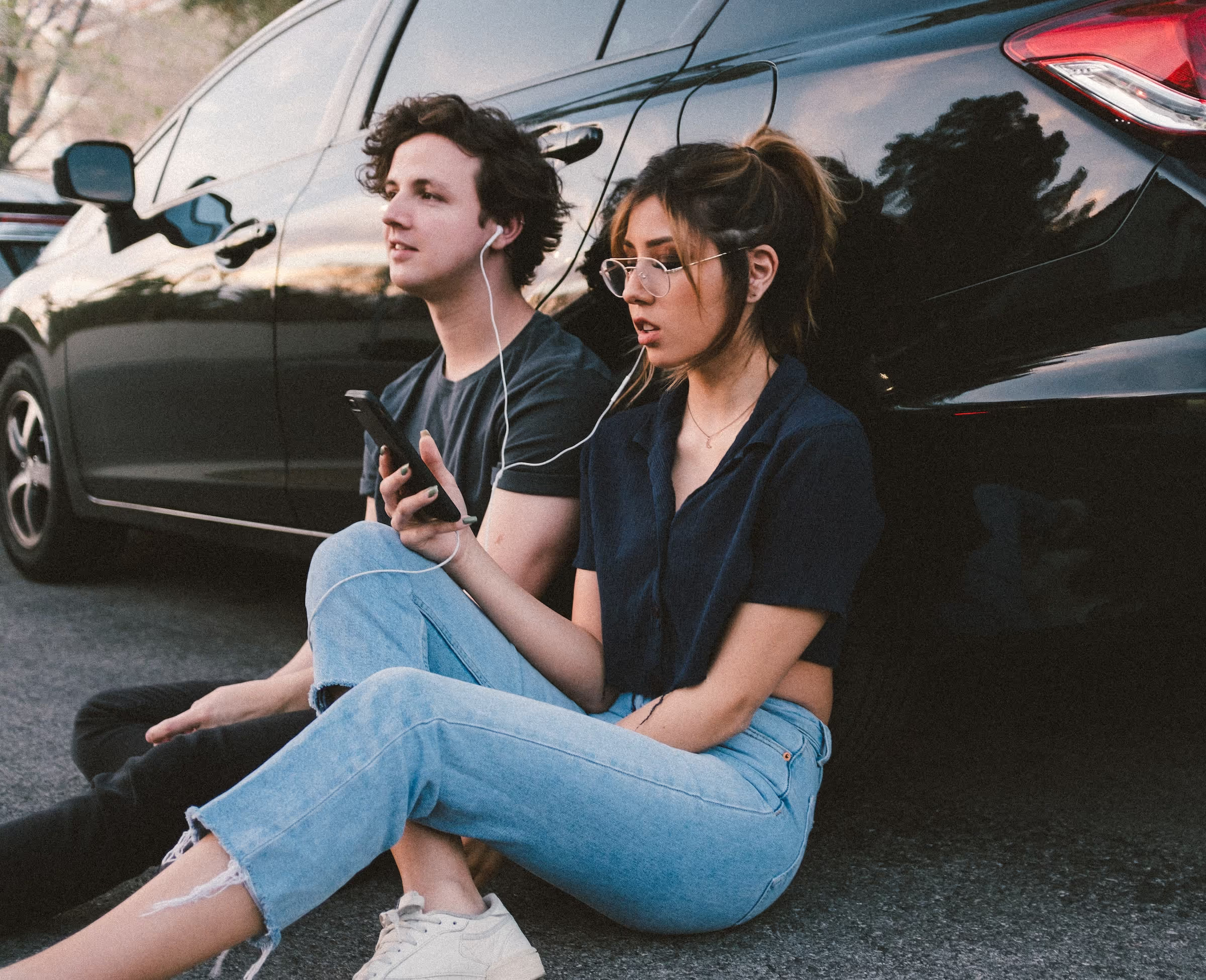 A young man and woman sit on the ground and lean against a parked car. They sahre a pair of wired earbuds and listen to music from a smart phone.